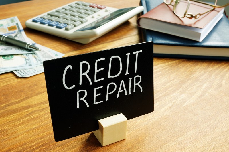 This image shows the top of a wooden desk with calculator, a stack of brown and blue books, some 0 bills, a pen, and a small chalkboard sign with the words "Credit Repair" written on it, representing the best credit repair affiliate programs.