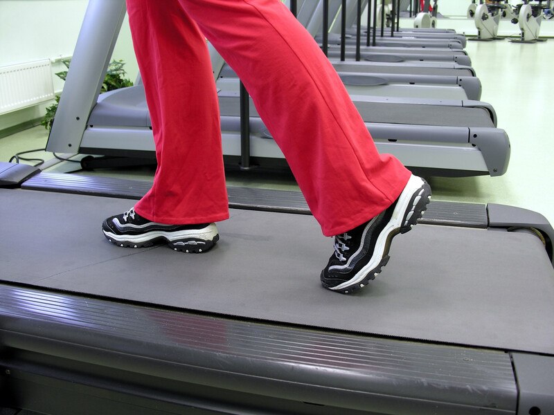 This image shows a row of gray treadmills, as well as the legs and feet of someone in red pants and black and white sneakers walking on the nearest treadmill, representing the best exercise machine affiliate programs.