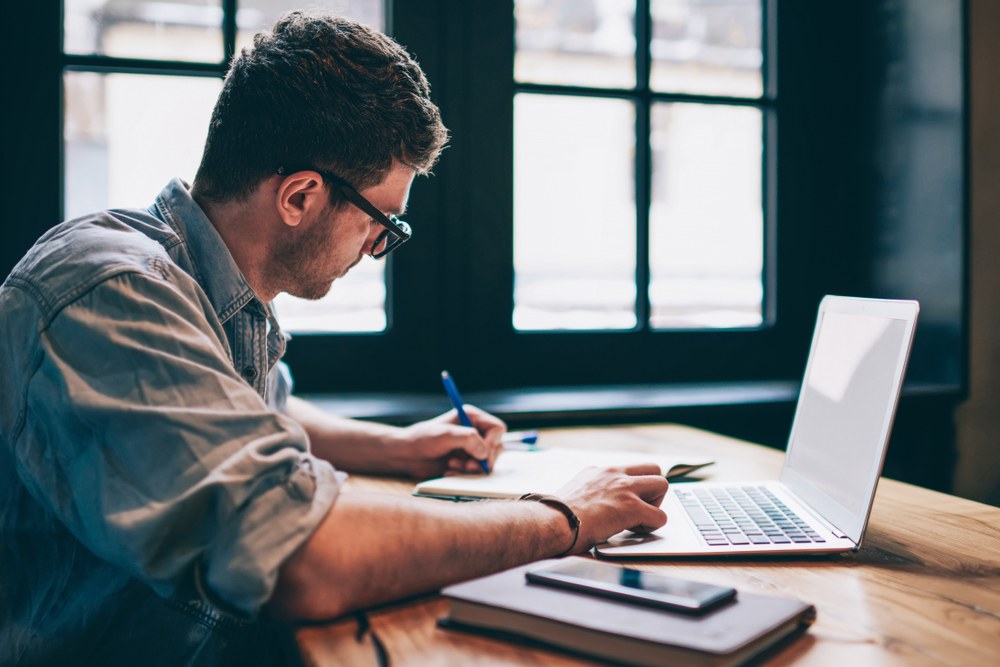 A young man sitting at a desk working on paper and a laptop