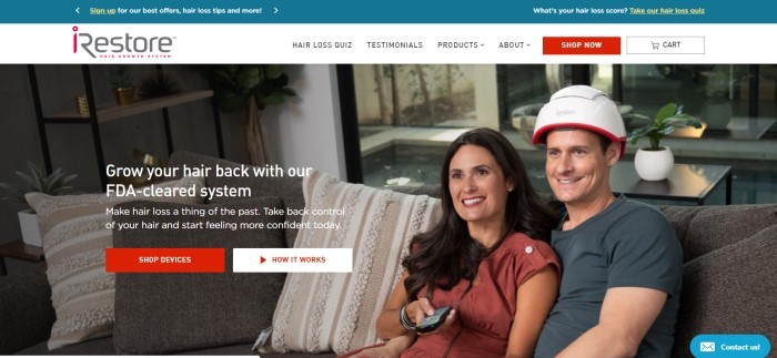 This screenshot of the home page for iRestore Laser shows a smiling woman with brown hair and a russet shirt holding a television remote control in her hand, while she snuggles next to a smiling man in a charcoal-colored tee shirt and an iRestore helmet on a brown and white couch. 