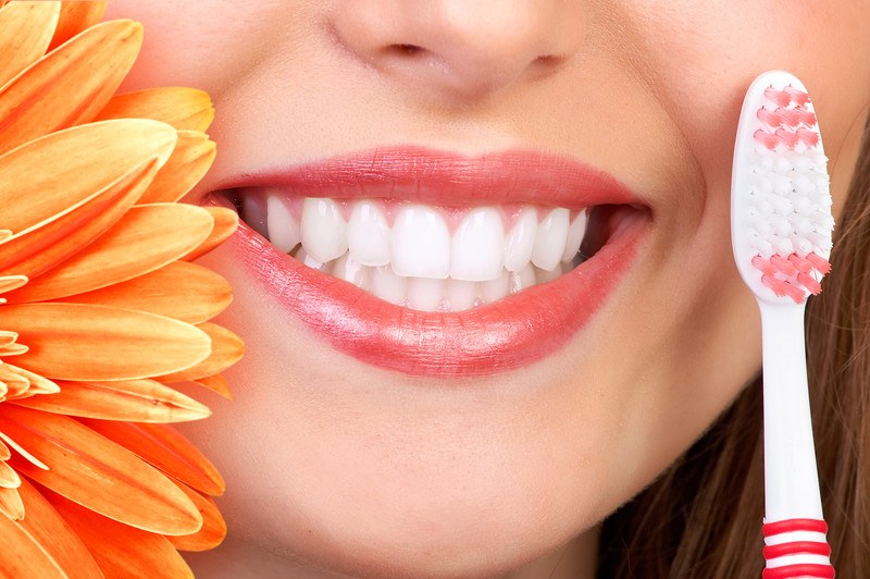 This photo shows the lower half of a smiling woman's face, along with petals of an orange flower and a red and white toothbrush, representing the best dental insurance affiliate programs.