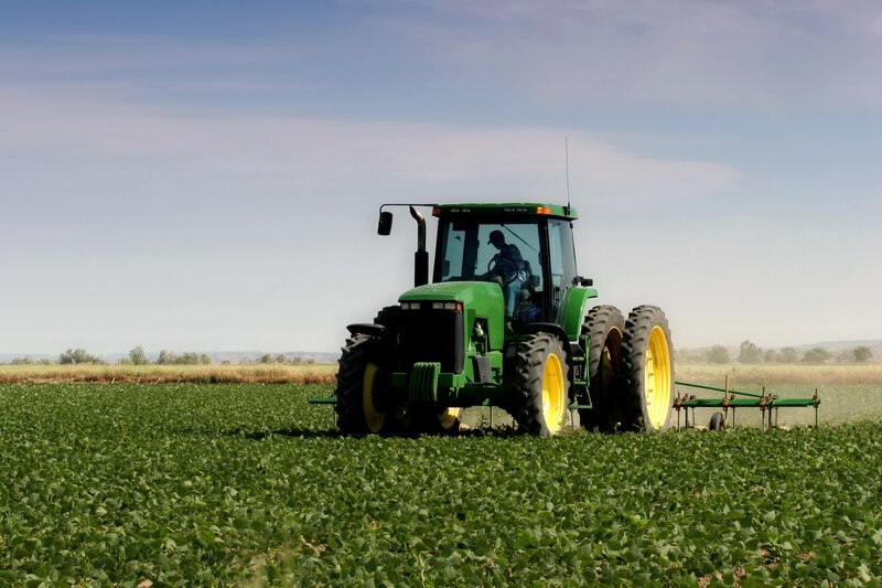 This photo shows a large green tractor with yellow and wheels and black tires pulling some farming equipment through a green field, representing the best farming affiliate programs.