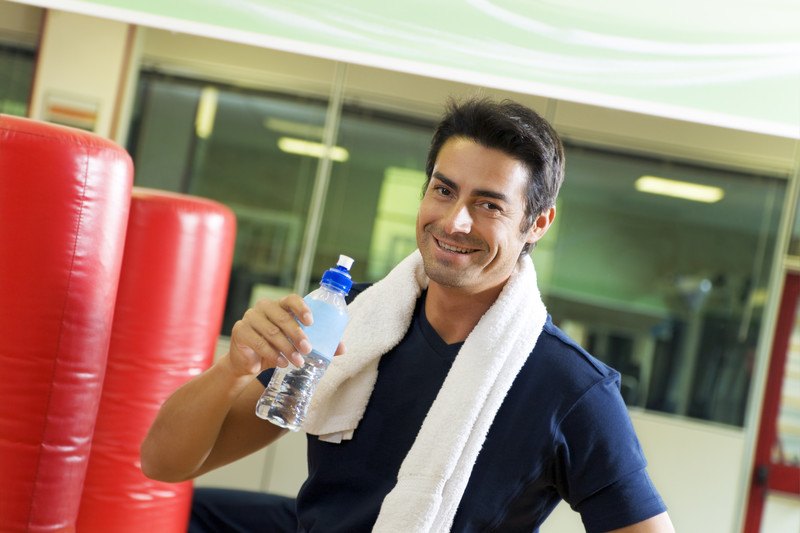 This image shows a smiling dark haired man in a blue tee shirt with a white towel draped around his neck, standing in a gym near some red kicking targets, holding a water bottle in his hand, representing the best men's health affiliate programs.