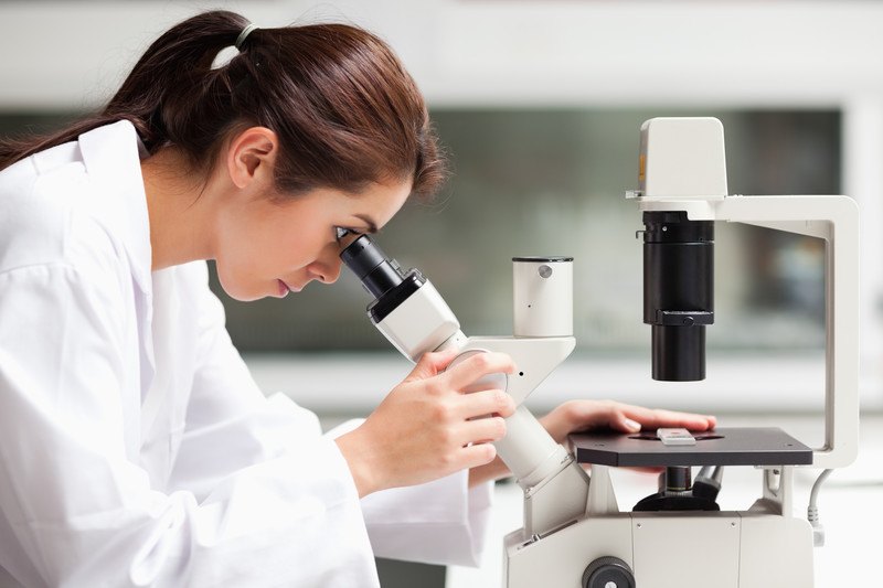 This image shows a dark-haired woman in a ponytail and a white lab coat looking into a white telescope in a laboratory, representing the best science affiliate programs.