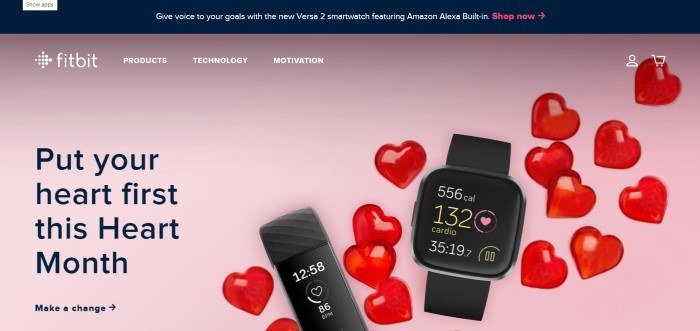 This screenshot of the home page for Fitbit has a black header announcing the new Versa 2 smartwatch above a photo with a pink background, red hearts, and two devices with heart monitors, along with black text inviting customers to put their hearts first.