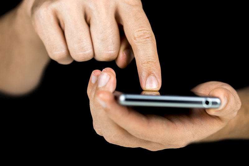 This photo shows a pair of male hands holding and tapping on a cell phone against a dark background, representing the best cell phone affiliate programs.