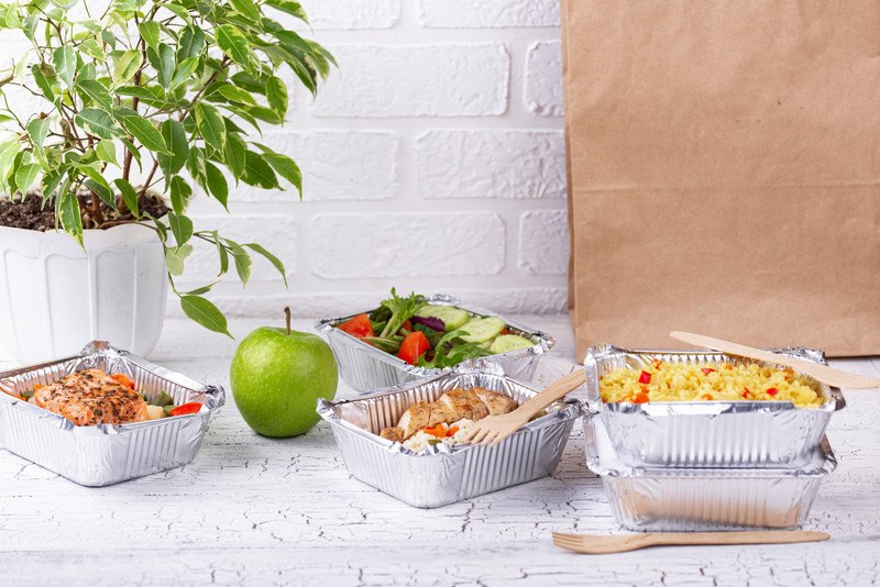 This photo shows four trays of food in foil containers sitting on a white table, along with a green apple, a house plant, and a brown paper bag, in front of a white brick wall, representing the best food delivery app affiliate programs.