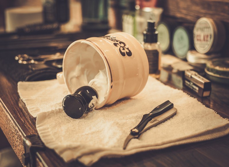 This photo shows a cream-colored shaving mug turned on its side, showing the shaving cream and a dark-handled shaving brush, lying on a white towel next to a pair of clippers on what appears to be a man's dresser, representing the best men's grooming affiliate programs.