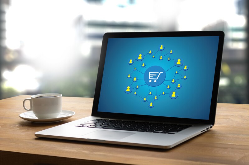 This photo shows an open laptop on a wooden desk next to a white teacup and saucer, and the laptop screen is blue with a shopping cart icon in the center and several yellow icons indicating shoppers surrounding it, representing the best online shopping affiliate programs for coronavirus.