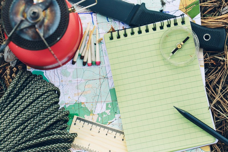 This photo shows an overhead view of survivalist supplies, including rope, matches, a map, a pen and notepad, a compass, and a knife, representing the best survivalist affiliate programs.