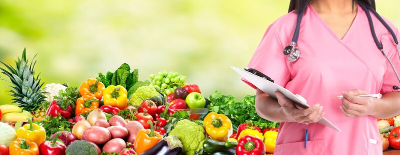 This picture shows a neck-down image of a woman in pink scrubs and a stethoscope, holding a clipboard while standing in front of a table full of brightly-colored fruits and vegetables, representing the best women's health affiliate programs.