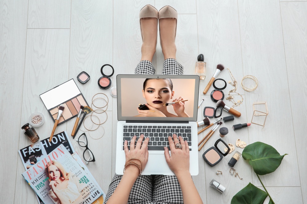 beauty blogger typing on mac keyboard with makeup products and fashion magazines in the background