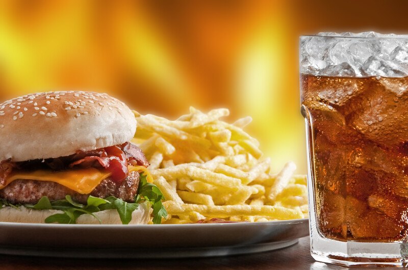 This image shows a cheeseburger and fries on a white plate, next to a cola drink in a clear glass, on a table in front of an orange background, representing the best fast food affiliate programs.