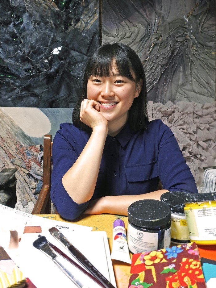 This photo shows a beautiful smiling Asian woman in a blue shirt sitting behind a table full of art supplies and in front of several finished textile art collages in grays and pinks, representing the best fine art affiliate programs.