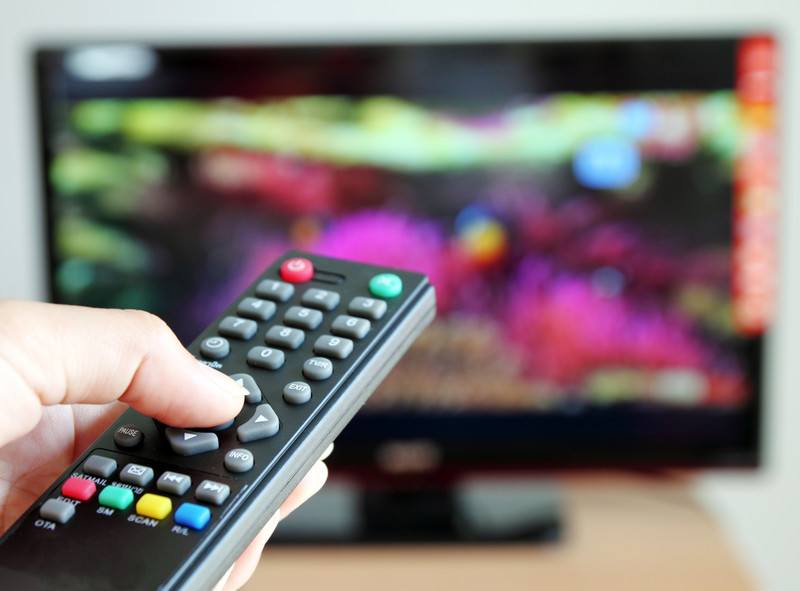 This photo shows a man's hand holding a television remote control, aimed at a blurred but colorful television screen, representing the best internet TV affiliate programs.