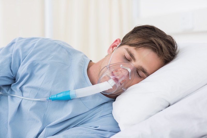 This photo shows a brunette man in a blue hospital gown lying with his eyes closed on a hospital bed, while he breathes through an oxygen mask, representing the best medical equipment affiliate programs.