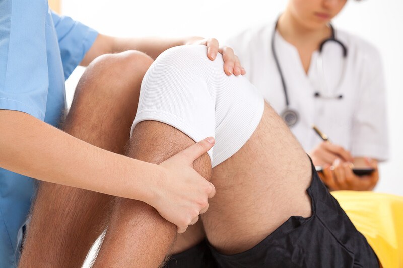 This photo shows the knees of a man in black workout shorts and a white knee brace as he is lying down near medical professionals in blue and white clothing, representing the best sports injury recovery affiliate programs.