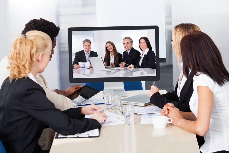 This photo shows a group of four people in business clothing gathered around a beige worktable, looking up at a screen showing four more business people in a different room, representing the best video conferencing affiliate programs.