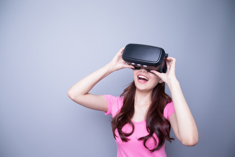 This photo shows a smiling woman with long brown hair and a pink shirt holding a set of virtual reality glasses up to her eyes in front of a gray background, representing the best virtual reality affiliate programs.