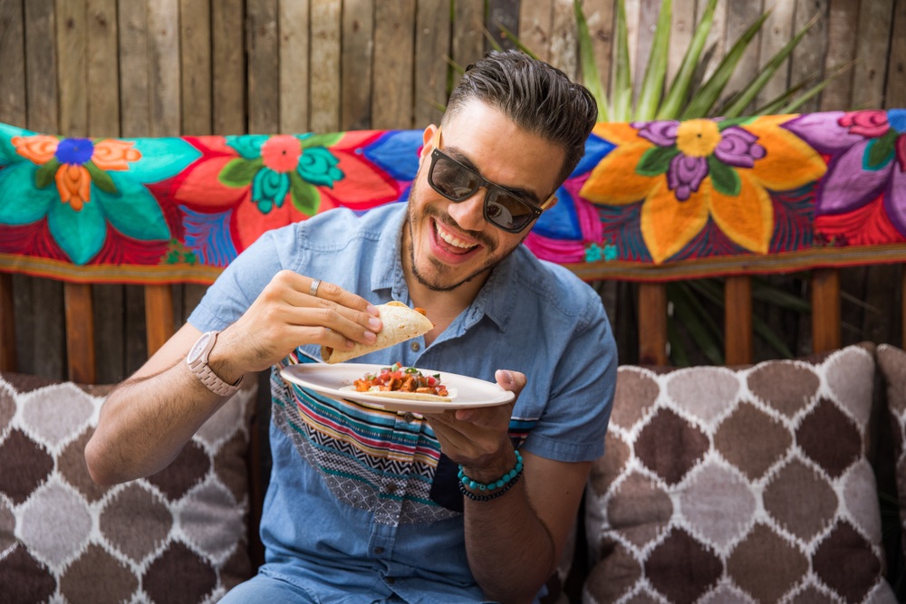 man with modern sunglasses and dark hair eating a taco with brightly colored material in background