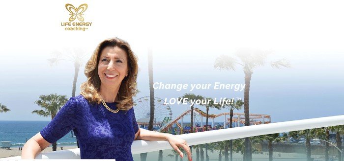 This screenshot of the home page for Life Energy Coaching has a gold logo above a photo of a smiling brunette woman in a blue shirt leaning against a balcony that overlooks a beachfront amusement park, along with white text inviting customers to change their energy and love their lives.