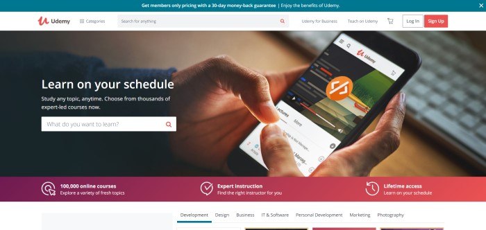 Alt: This screenshot of the home page for Udemy shows a photograph of a man's hands accessing Udemy classes on a mobile phone, behind white text inviting potential students to learn on their own schedule. Caption: Udemy is a marketplace for purchasing courses and learning in a self-paced, online environment.