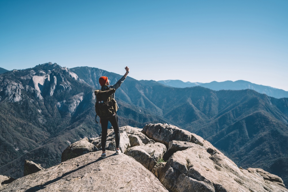 woman with backpack standing on top fo a rock with huge mountains in the background