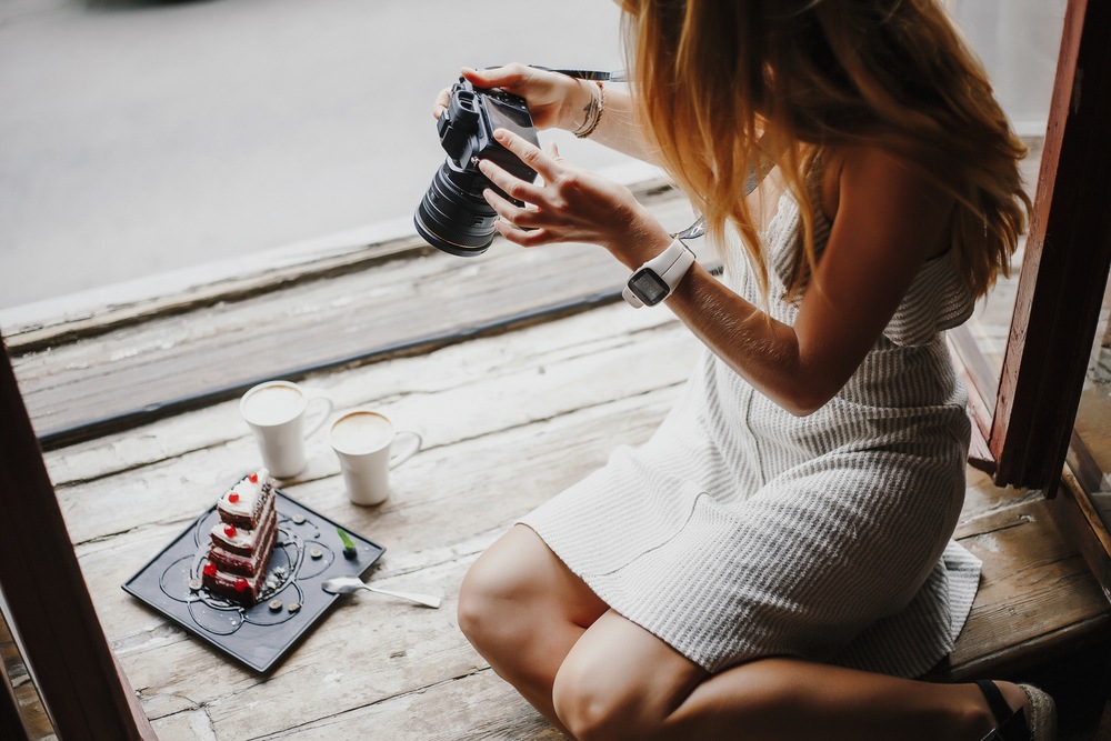 young woman taking professional photos of layered cake with coffee sitting on wooden window sill