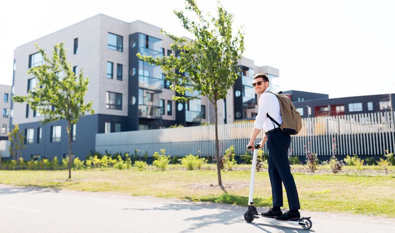 This photo shows a young man in sunglasses, a white shirt, and dark pants riding on an electric scooter with a brown backpack on his back on a walking path near an office building, representing the best scooter affiliate programs.