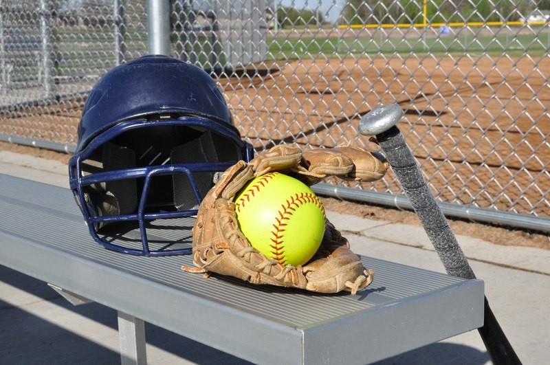 This photo shows a dark blue helmet, a brown mitt, a black bat, and a yellow softball on or near an aluminum bench in front of a chain-link fence near a softball field, representing the best softball affiliate programs.