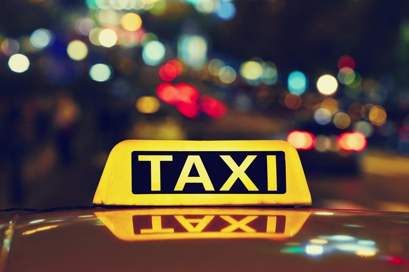 This photo shows the a yellow and black light bar reading 'Taxi' on the top of a car in front of blurry city lights in red, blue, green, orange, and white against a dark sky, representing the best taxi affiliate programs.