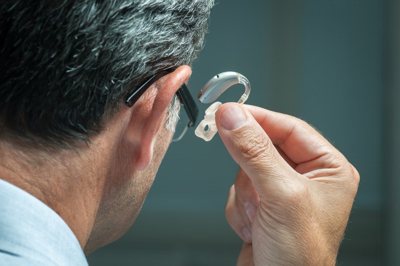 This photo shows a dark-haired man in a blue collared shirt and black glasses lifting a silver hearing aid to his ear, representing the best hearing aid affiliate programs.