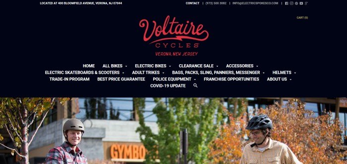 This screenshot of the home page for Electric Spokes has a black background, an announcement in red text for Voltaire Cycles, white text in the black navigation bar, and a photo of two smiling men in helmets and plaid shirts preparing to ride on electric scooters outside what appears to be an office building or shopping center.