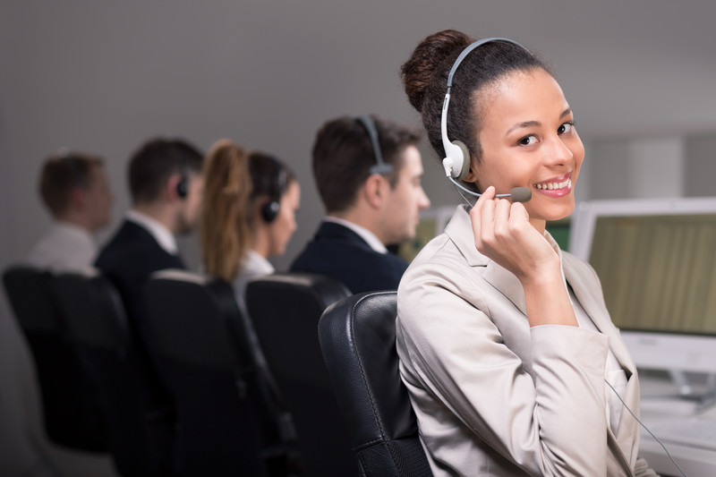 This photo shows a smiling woman in light-colored business clothing and a headset looking at the camera at the end of a row of similarly-dressed people who are looking at computer screens in front of them, representing the best call center affiliate programs.