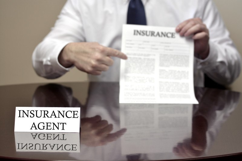 This photo shows the shoulders and arms of a businessman in a white suit and dark tie holding a white paper with black text and the word 'Insurance' at the top above a polished wooden desk with an insurance placard on top of it, leading readers to ask: do insurance agents make good money?