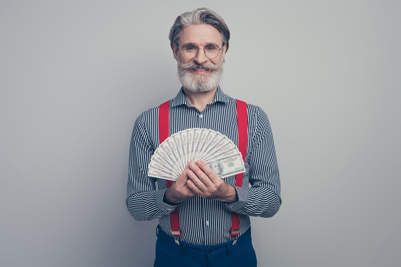 This photo shows a smiling white-bearded man in a striped shirt, blue pants, and red suspenders holding several 0 bills in a fan shape in front of him, representing the best lifetime commissions affiliate programs.