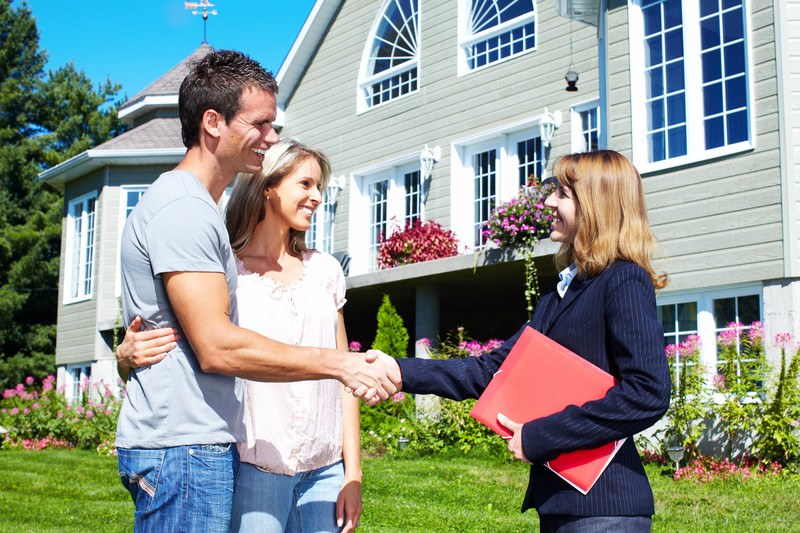 This photo shows a smiling woman real estate agent in a dark blue jacket, holding a red folder, shaking hands with a smiling couple on the lawn in front of a large gray and white house.