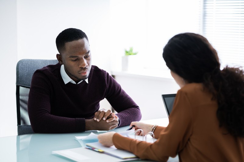 This photo shows a dark-skinned, dark-haired man and woman in business clothing discussing financial documents across a blue table, representing the best tax preparers in action.