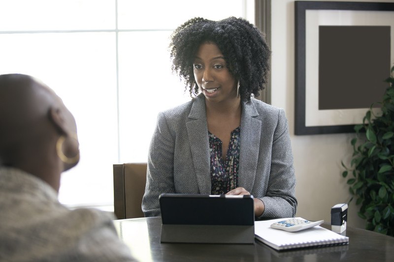 This photo shows a dark, curly-haired woman in a gray business jacket sitting in front of a window, with a computer in front of her, talking with another woman about finances, representing the best tax preparers