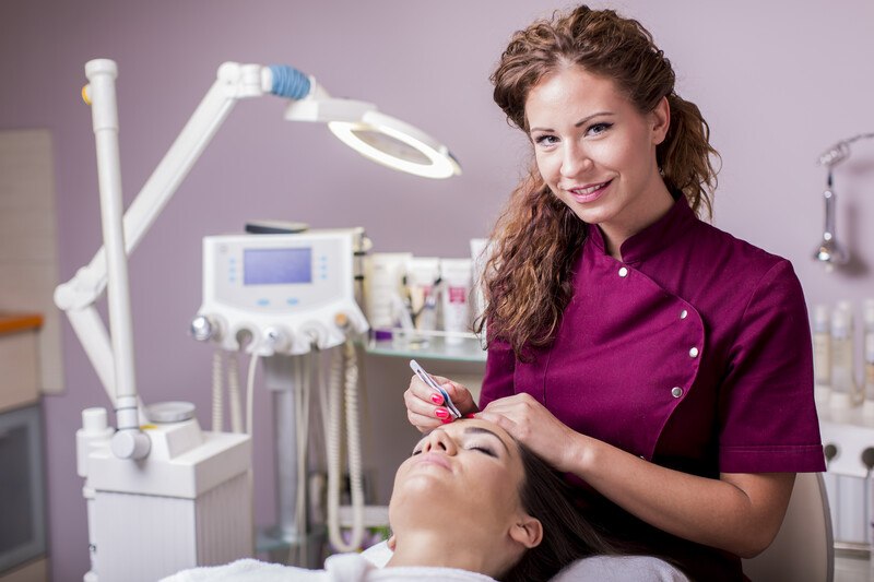 This photo shows a smiling brunette esthetician in a burgundy lab coat doing a cosmetic procedure on a woman lying on the table in front of her, representing an answer to the question: Do estheticians make good money?