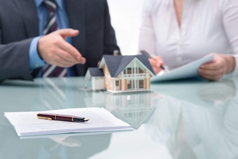 This photo shows the shoulders of a man in a business suit, blue shirt and striped tie next to a woman in a white shirt who is writing something on a piece of paper while the ma gestures toward a model of a pink and white house near a stack of paperwork and a pen lying on a shiny table in front of them.