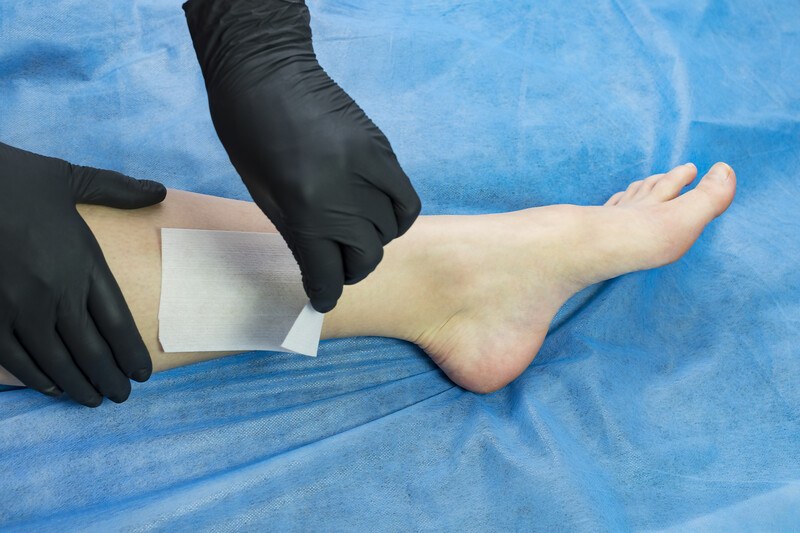 This photo shows a pair of hands in black gloves peeling wax from a woman's legs, on top of a blue blanket.