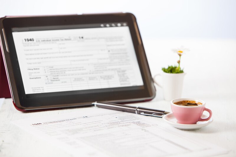 This photo shows a tax document 1040 loaded on the screen of a small computer, along with a potted plant, a pen, printed tax forms, and a pink coffee cup with coffee in it on a white saucer.