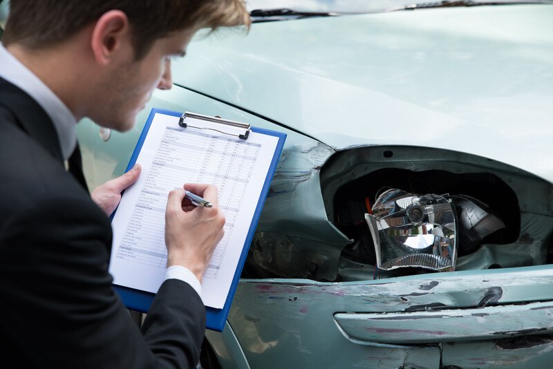 This photo shows a brown-haired male insurance agent writing something a clipboard as he looks at damage done to a light-colored car near one of the headlights.