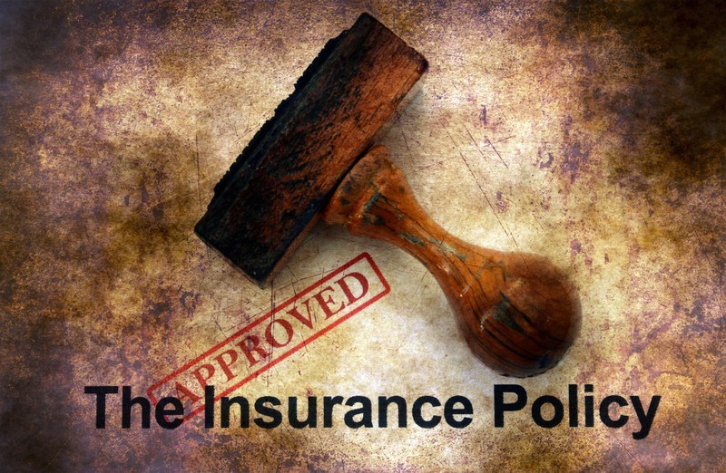 This photo shows a wooden stamp and a red stamp of approval on an insurance policy on a grown and white background.