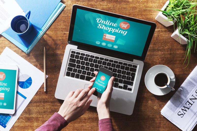 This overhead photo shows a woman's hands holding a mobile phone above a laptop, both of which have an online shopping screen loaded, along with papers, coffee cups, and potted plants on wooden desk, representing the best online retail affiliate programs.