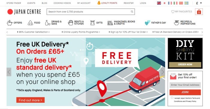 This screenshot of the home page for Japan Centre has a white background with black text and navigation bar above a graphic of a red bus driving on a gray map with red text announcing free delivery, with a blue text section on the left with black and red text and a red call to action button and an advertising column on the right.