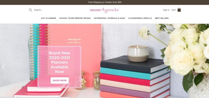 This screenshot of the home page for MomAgenda has a dark header, a white navigation bar with a pink logo, and a photo showing a variety of different day planners and organizer books in pink, aqua, black, purple, and beige on a whte table next to a beige candle jar with a lit candle and a vase of white flowers.