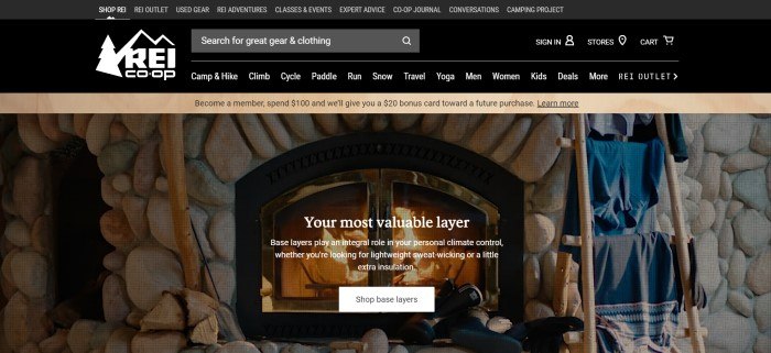 This screenshot of the home page for REI has a black header and navigation bar above a photo of a lit fire inside a rock fireplace, along with clothing racks on the right side where jackets in blue and gray are hanging.