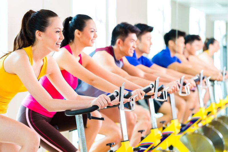 This photo shows a row of smiling dark-haired men and women in various brightly colored fitness clothing, working out on a row of yellow exercise bikes, representing the best fitness clothing affiliate program.